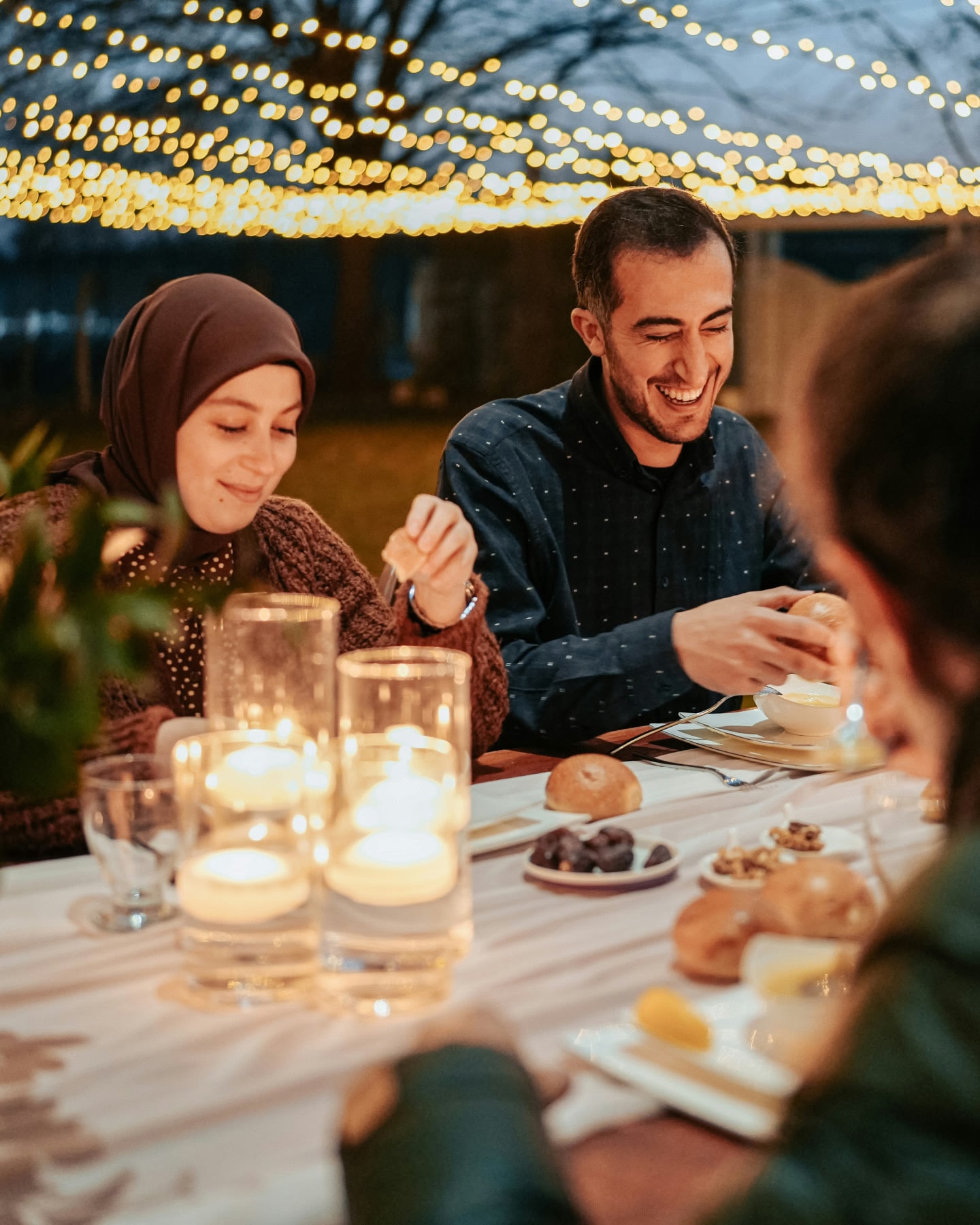 أصدقاء يتشاركون وجبة على ضوء الشموع، تتدلى فوقهم أضواء متوهجة.