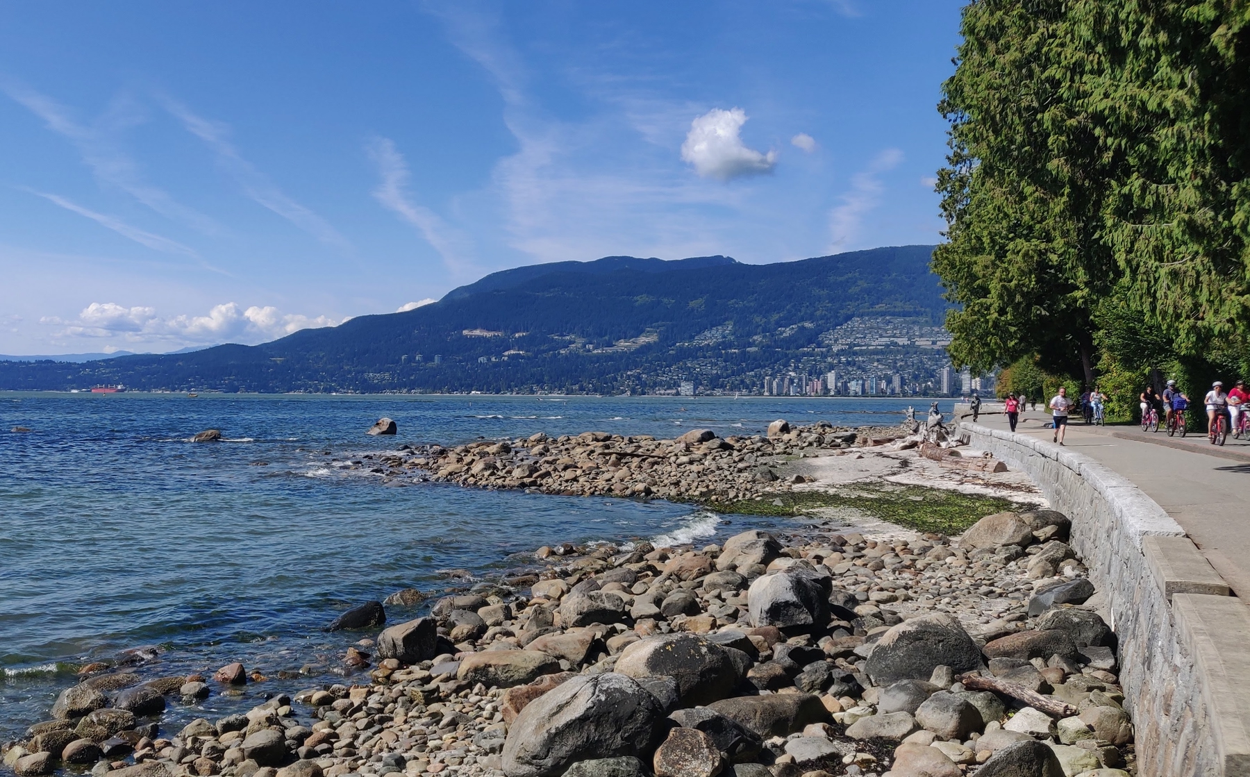 rocks by the shore and people bicycling on the seawall in Vancouver by Stanley Park. Rocky Mountains in the background.