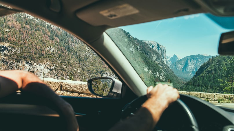 View from inside a car driving through a mountainous region. The driver's hands are on the steering wheel, and the landscape features trees, rocks, and a clear blue sky.
