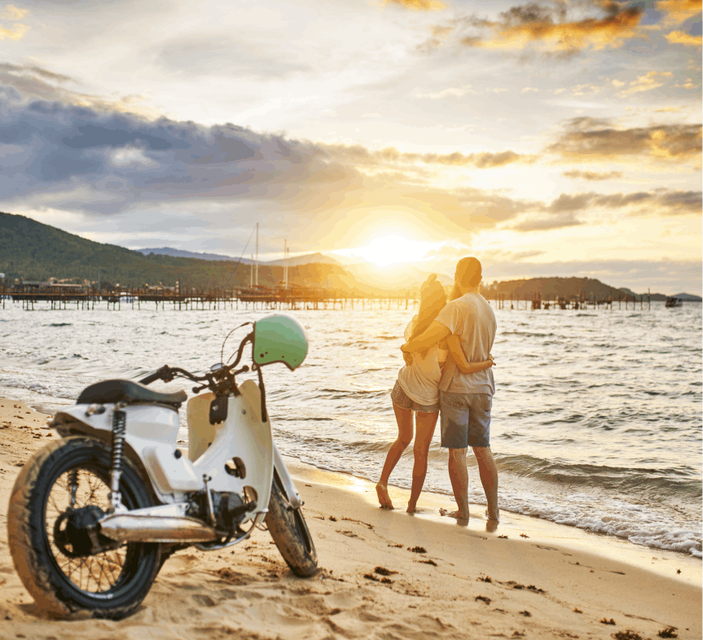 A parked motorbike on a picturesque beach, with the serene sea in the background. A romantic couple embraces in front of the bike. 