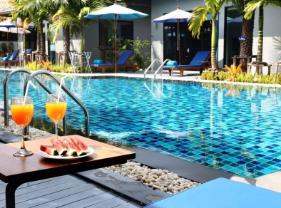 Eine erfrischende Aufnahme eines luxuriösen Hotelresort-Pools mit kristallklarem Wasser, das die Gäste zum Schwimmen und Abkühlen von der warmen Sonne einlädt. Im Vordergrund ist ein stilvoller Tisch mit zwei köstlich aussehenden Cocktails und einem Teller mit frischen Wassermelonenscheiben gedeckt, was zur entspannten und genussvollen Atmosphäre der Szene beiträgt.