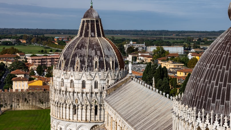Una vista del Battistero di Pisa in Italia, con la sua cupola ornata e l'architettura dettagliata, sullo sfondo campi verdi, alberi e edifici vicini.