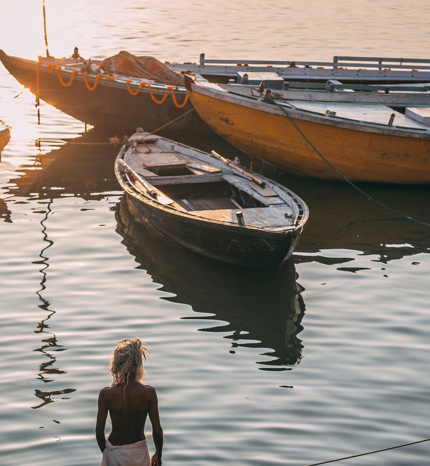 A person stands in water with their back to the camera next to a small boat as the sun sets in Varanasi, India.