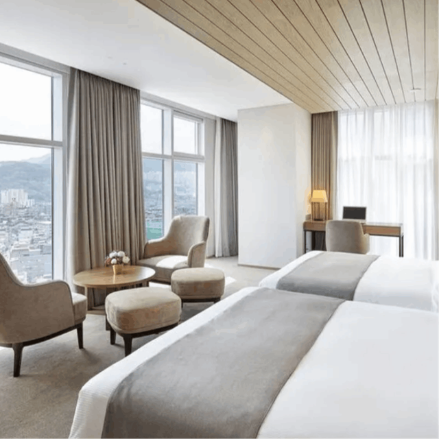 A glimpse inside a room at Staz Hotel Doksan, featuring two white beds and a window with a city view. Adjacent to the window are two chairs and a table, creating a comfortable and scenic setting.