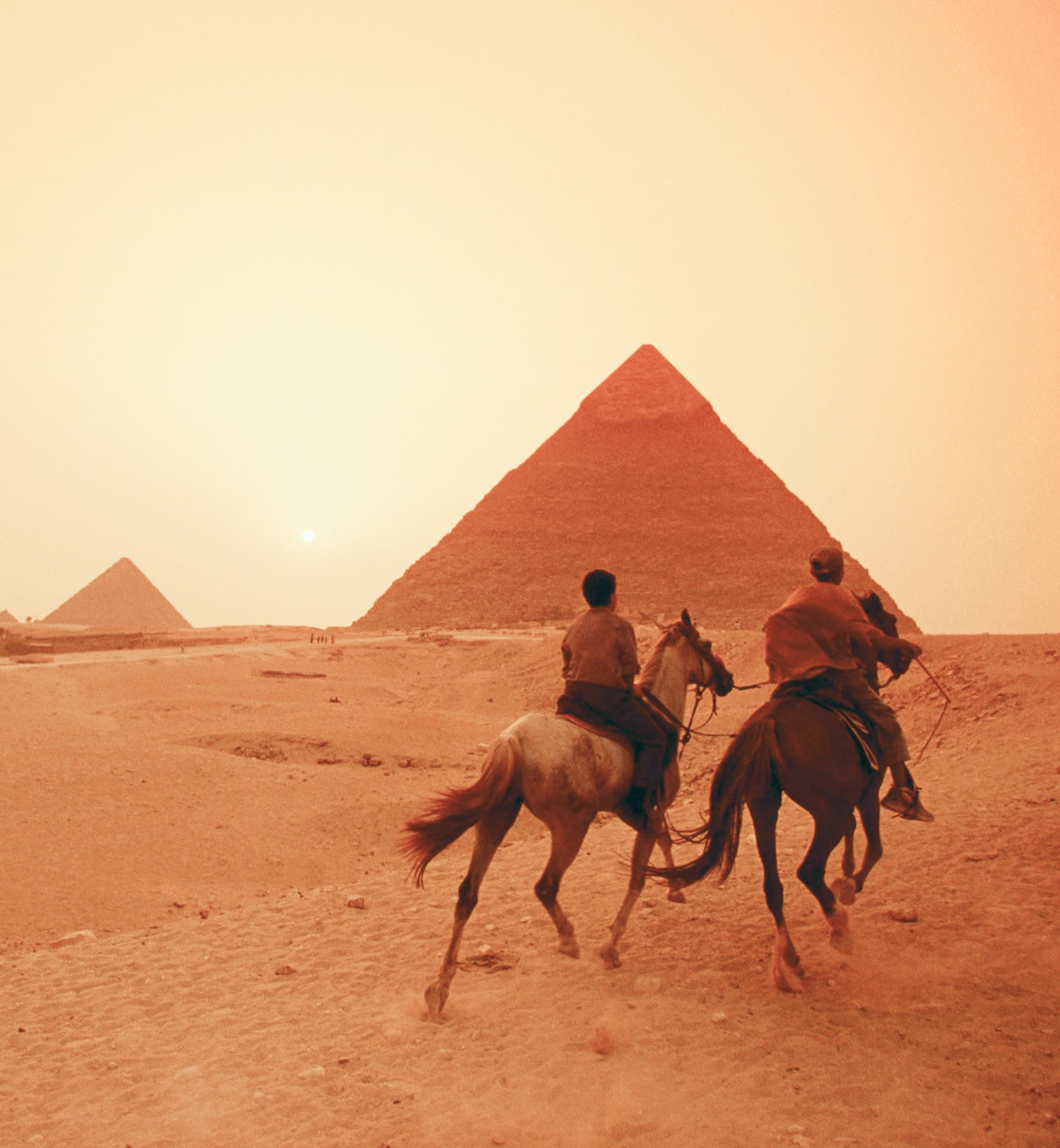 Zwei Reiter galoppieren auf die Pyramiden von Gizeh, Ägypten, bei einem warmen Sonnenuntergang in der Wüste zu.