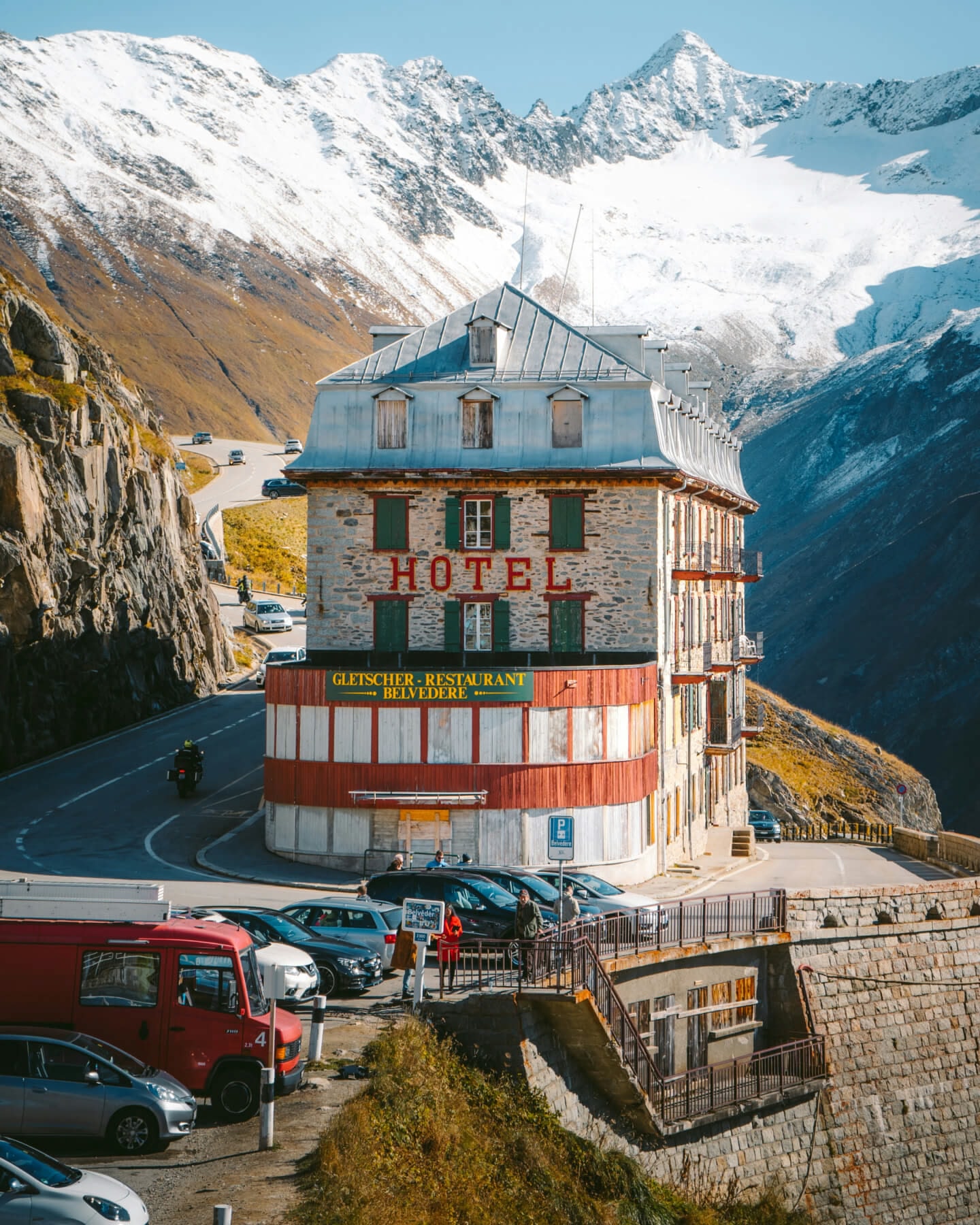 An Alpine-style hotel with a white, green and red façade, with mountains in the background