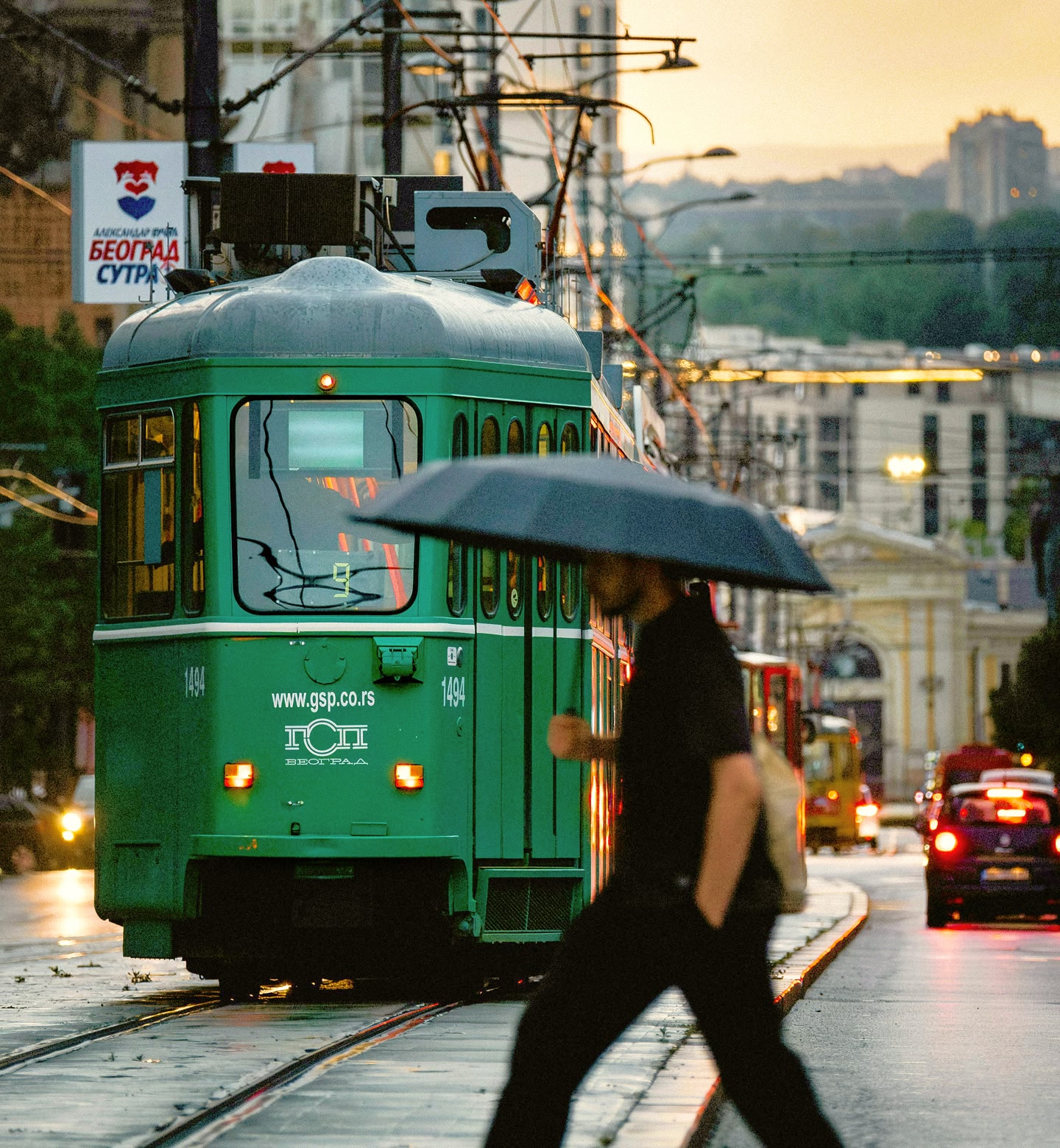 Ein Mann mit einem Regenschirm überquert die Gleise vor einer grünen Strassenbahn in Belgrad, Serbien.