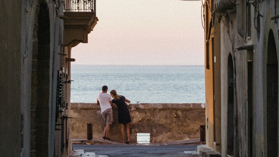 Una coppia vista di spalle si affaccia su un muretto che dà sul mare al tramonto, incorniciata da due edifici in una stretta via.