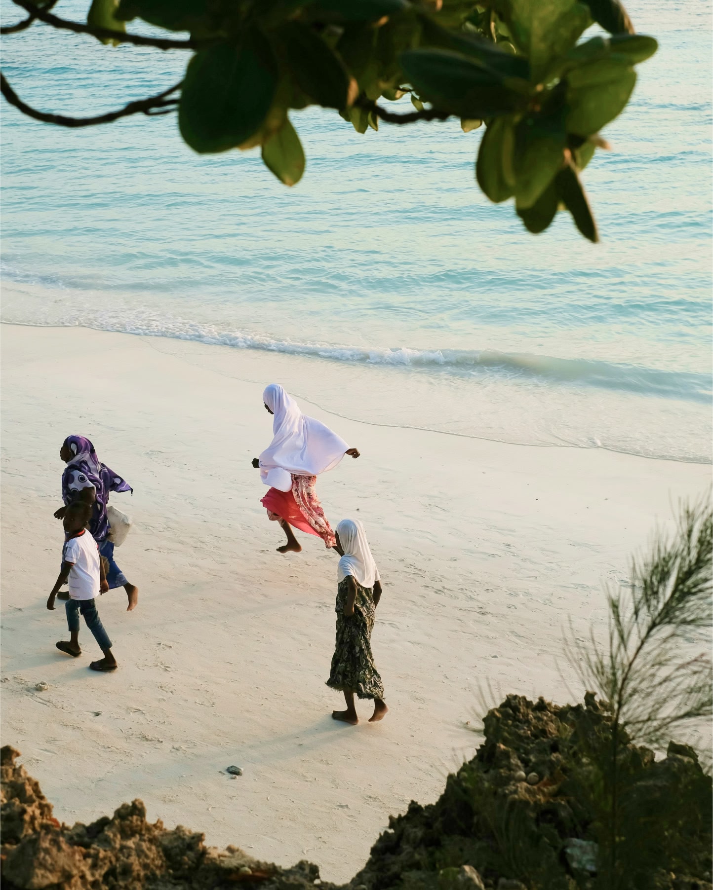 Three women wearing headscarves and a young boy run barefoot across a sandy beach.