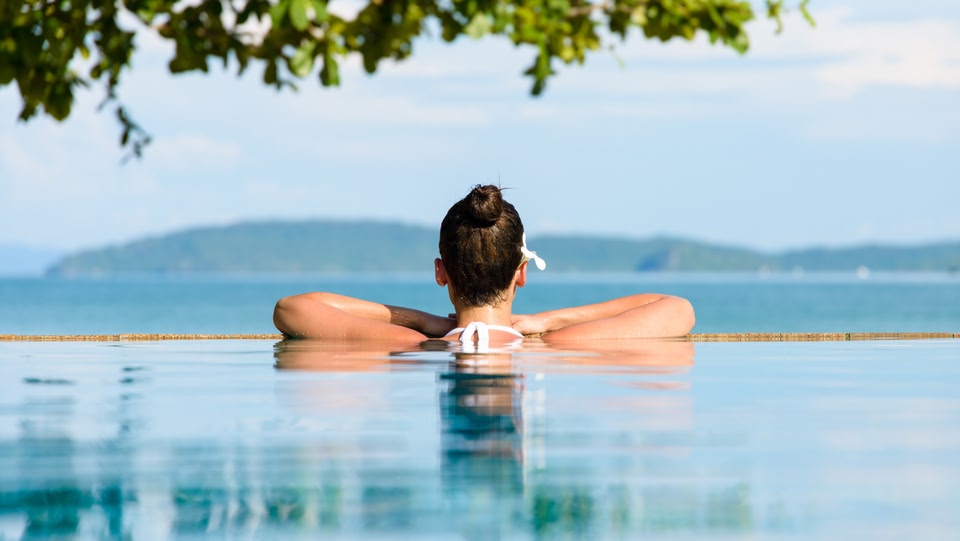 An image of a women in a pool with her shoulders out of the water, looking out into the ocean. 