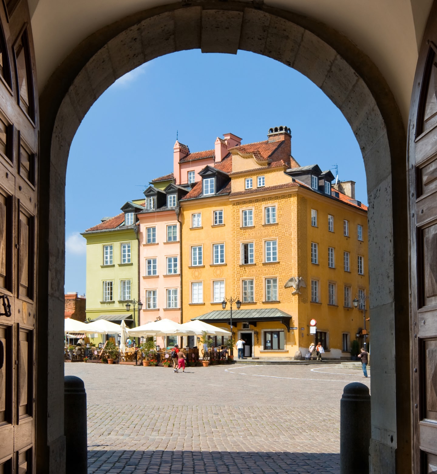 Tall green, pink, and yellow buildings with cafe umbrellas in front of it are viewed from a distant archway in Warsaw, Poland
