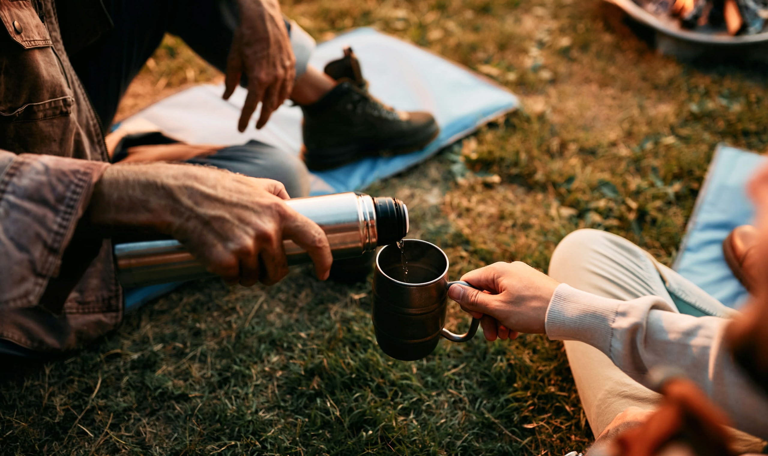 A person's hand pouring coffee from a flask into a mug held by another person as they sit on grass wearing hiking shoes