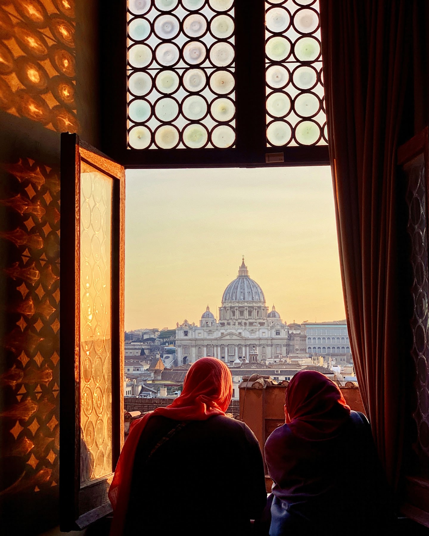 Two women wearing headscarves at a window looking out to St Peter's Basilica, Rome, at sunset