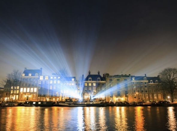 Canal in Amsterdam at night during Amsterdam Lights Festival. A number of lights beam up into the sky from the pavement.