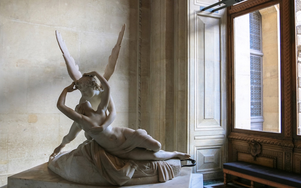 Marble statue in the Louvre, a Paris tourist attraction, featuring an angel cradling a woman. The statue is placed in a corner near an ornate window.