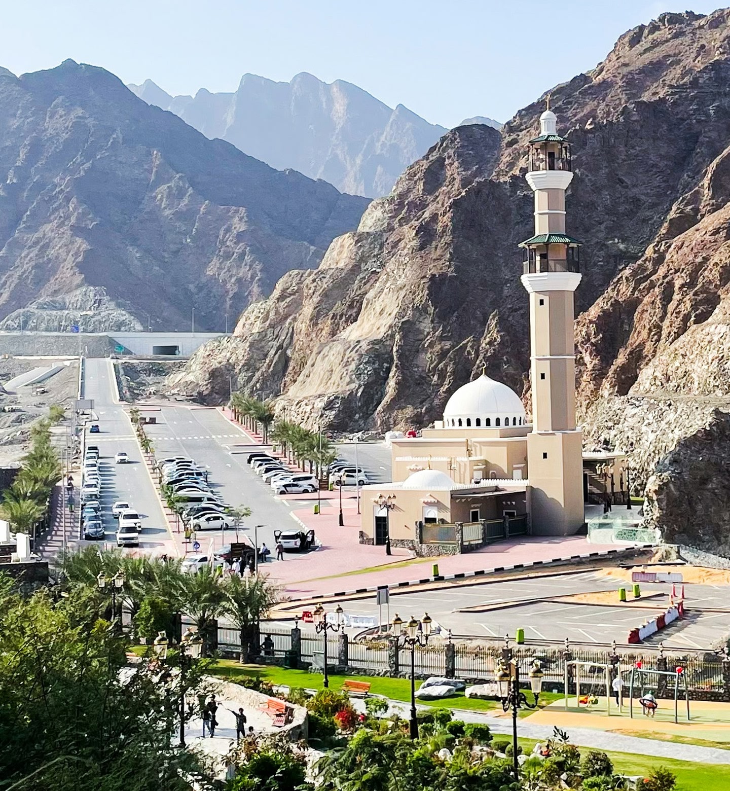 A mosque stands between rugged peaks and a busy car park in Sharjah, UAE.