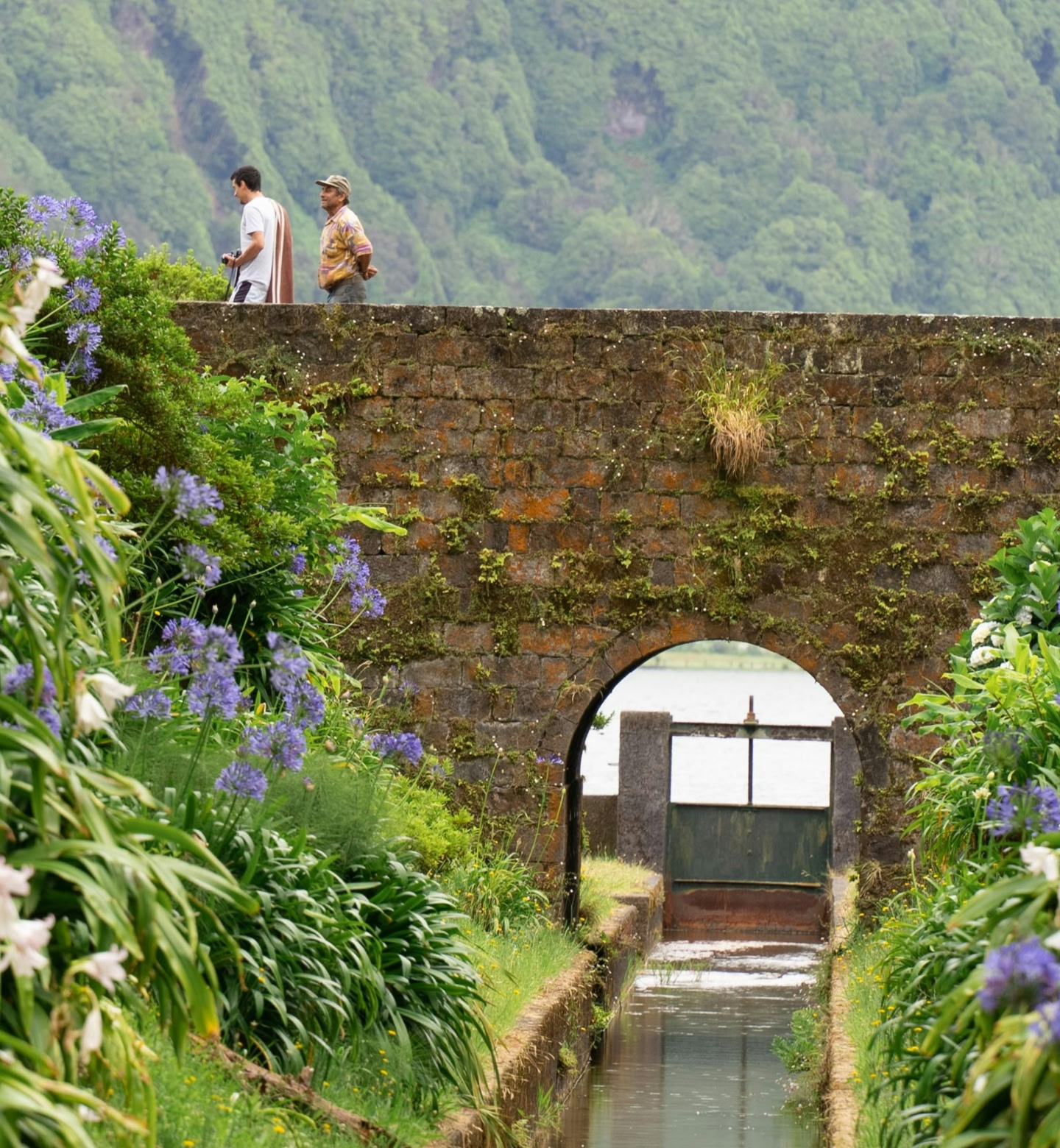 Two men cross an old brick bridge in a mountainous, verdant scene in rural Ponta Delgada, Portugal.