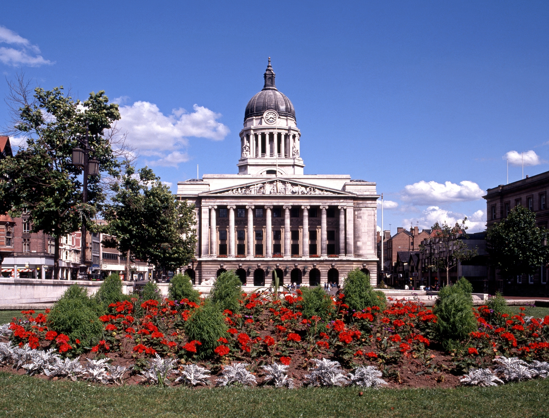 Picture shows Nottingham Council House with a colourful flower bed with green bushes and red flowers in front of it.