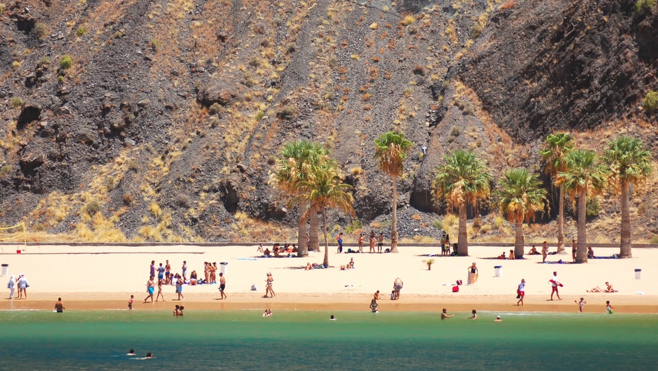 Crowded beach with palm trees and a rugged, rocky hillside as a backdrop.