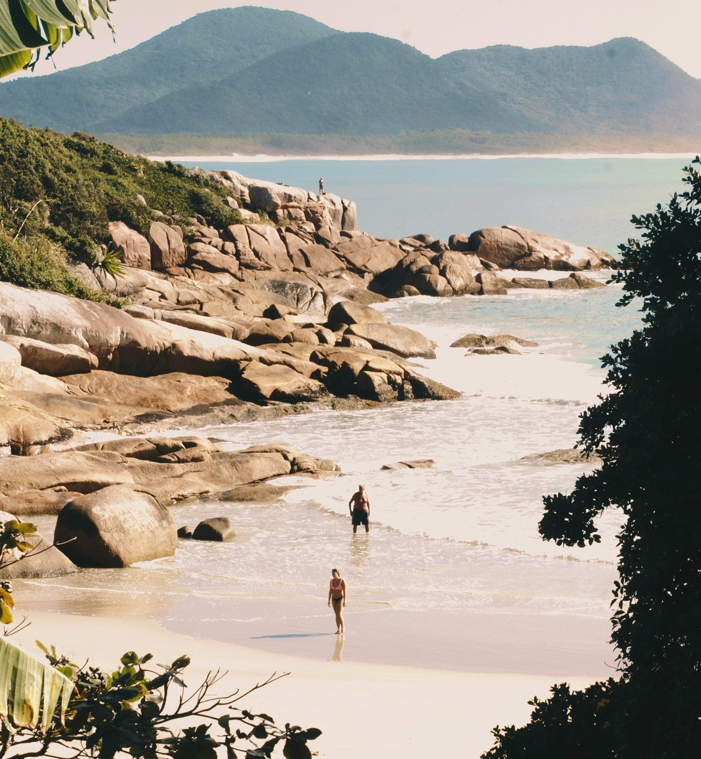 Two people stand in the sea beside a rocky outcrop on a beach in Victoria, Seychelles.