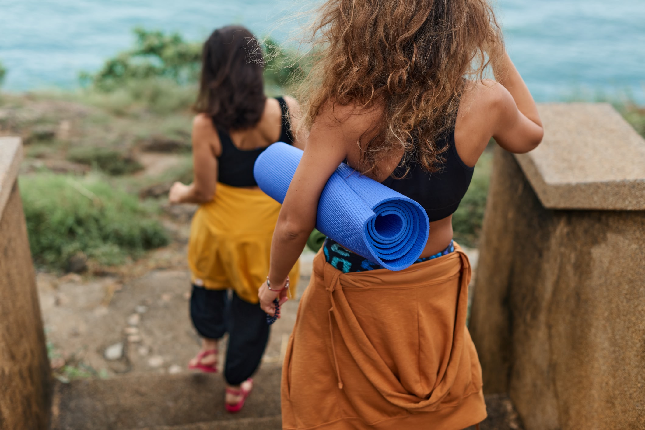 Young women holding exercise mat walking to cape to do yoga in nature, outdoors. View from behind.