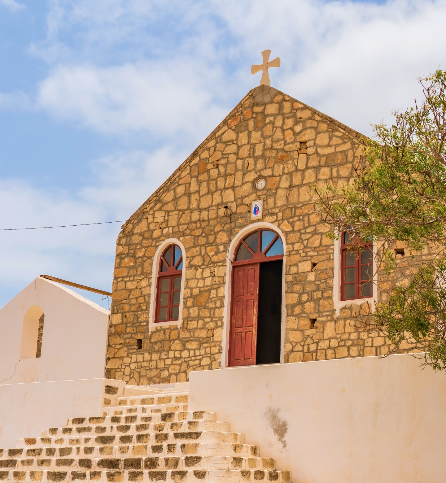 Une petite chapelle en pierre surmontée d'une croix et dotée d'une porte rouge, sous un ciel radieux à Maio, au Cap-Vert.