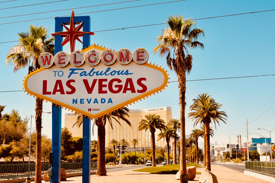The iconic "Welcome to Fabulous Las Vegas" neon sign stands on a sunny median lined with palm trees, with the Mandalay Bay hotel visible in the background.