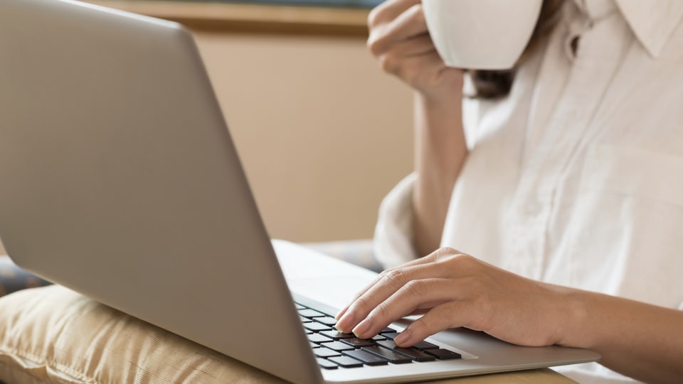 Image of a women sitting drinking out of a cup whilst typing with one hand on a silver laptop
