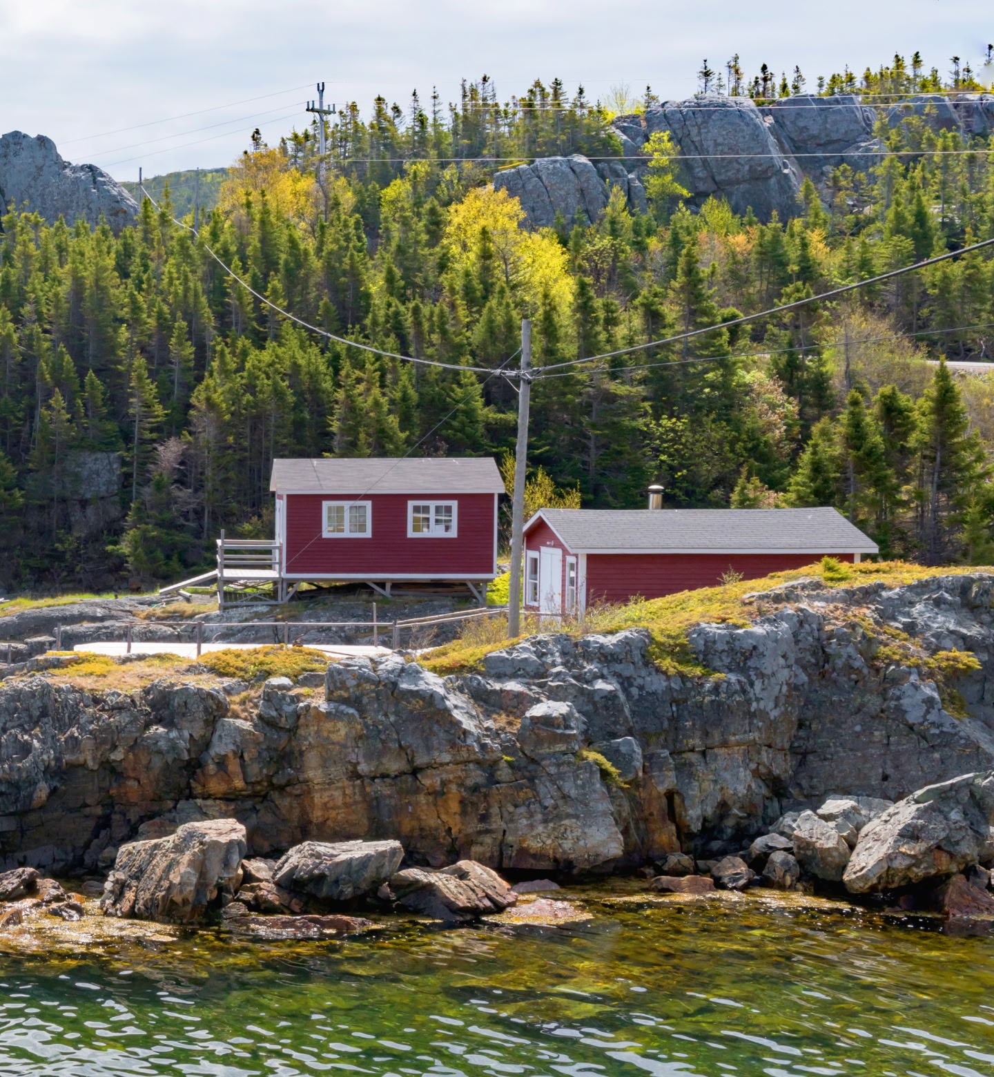 Two wooden homes sit astride the rockface on the coast of Gander, Newfoundland, with trees in the background.