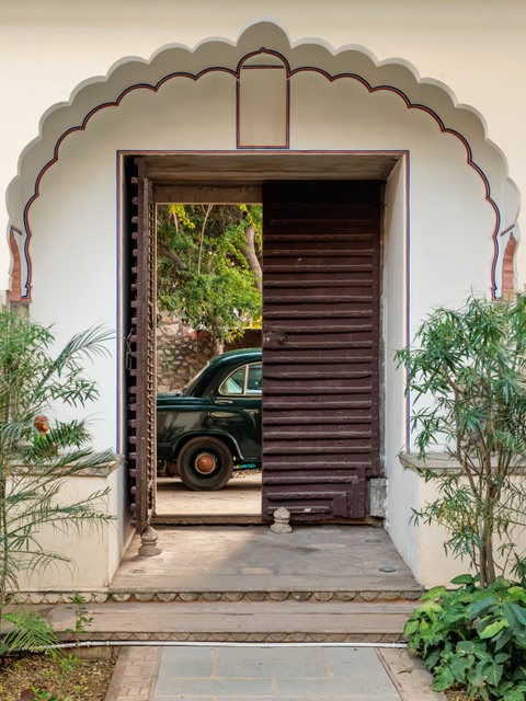 A vintage dark green Ambassador car glimpsed through a traditional scalloped arched wooden doorway at Dera Mandawa, Jaipur, framed by lush green garden foliage on either side of the white haveli entrance.