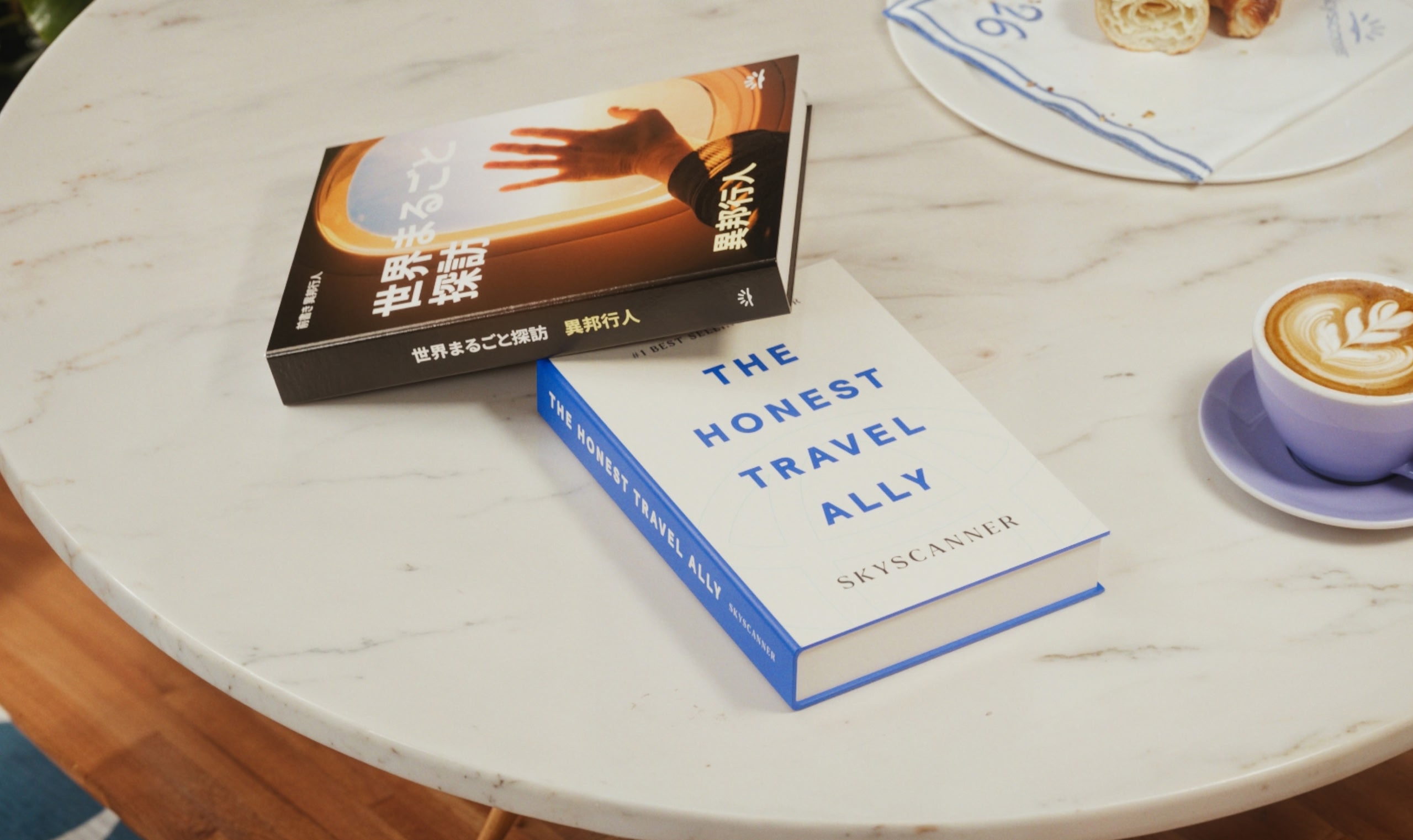Dos libros apilados sobre una mesa blanca junto a una taza azul de café y un plato con pastelitos