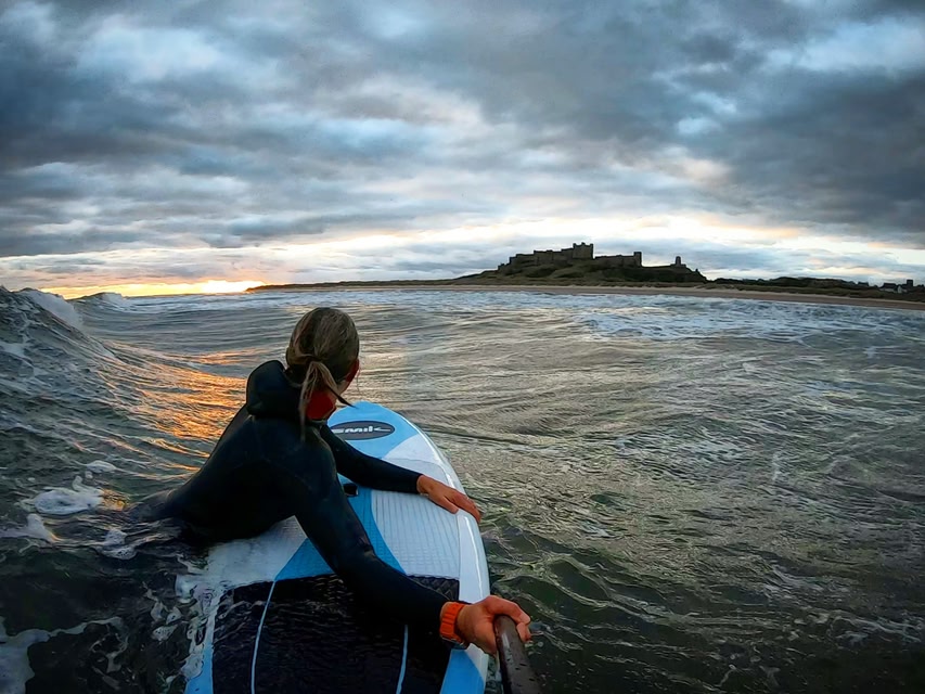 Rear view of a woman floating in the sea with paddleboard, looking towards shore and Bamburgh Castle, Northumberland, England.