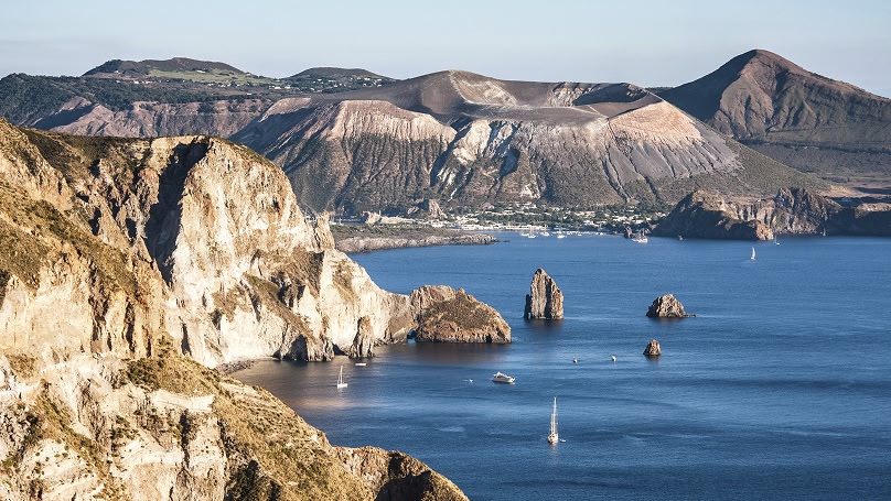 Panorama di un'isola vulcanica con scogliere, mare blu e montagne sullo sfondo.
