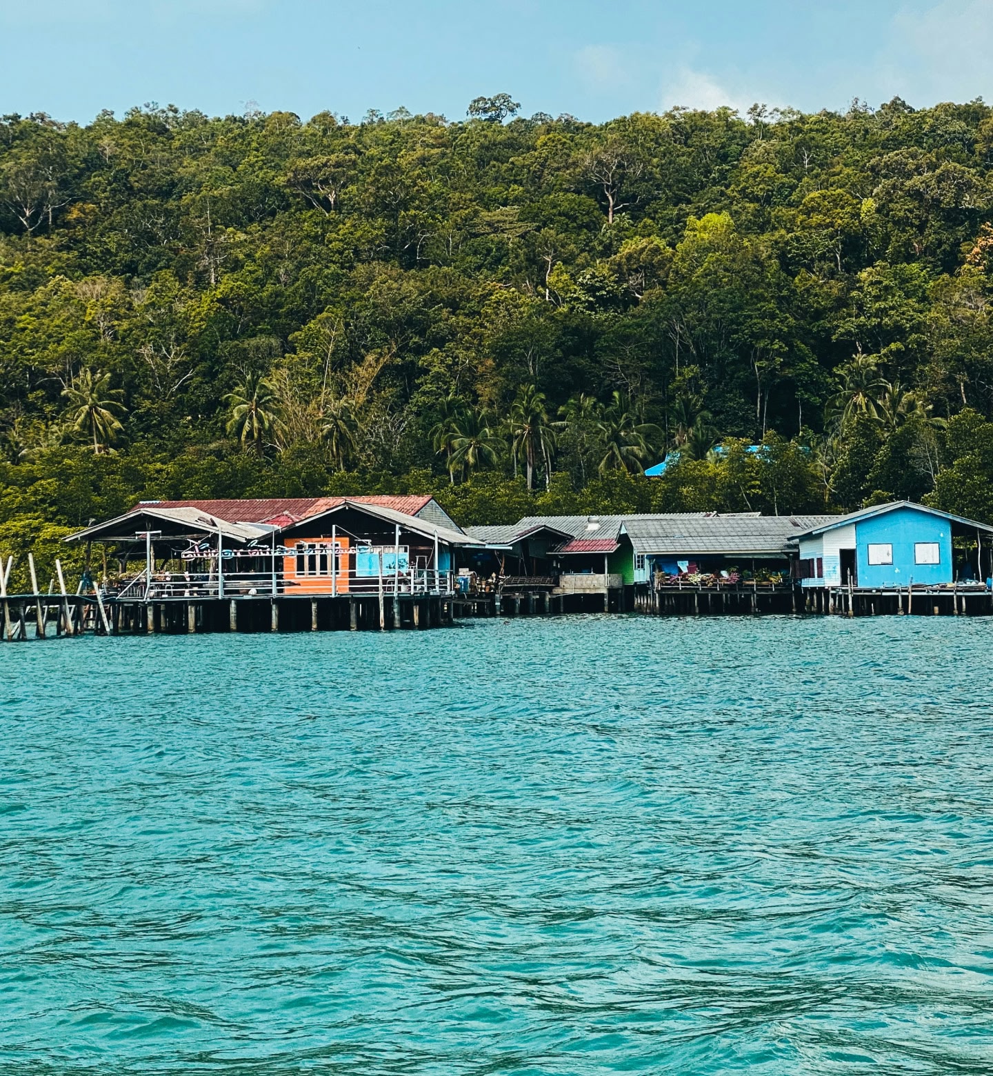 Brightly coloured houses alongside a coastline sit on stilts in the ocean, against a background of a rainforest in Luganville, Vanuatu