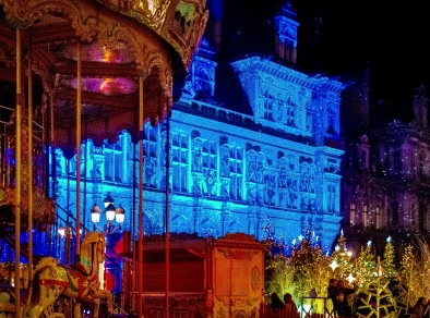 A picture of an historic building lit up with blue light on its façade with a row of Christmas trees in front of it.