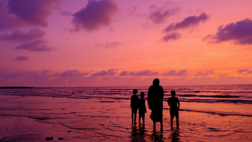 A serene family moment captured on a beach during dusk, with silhouettes of a woman and three young children gazing in awe at the breathtaking orange sky and tranquil sea ahead.