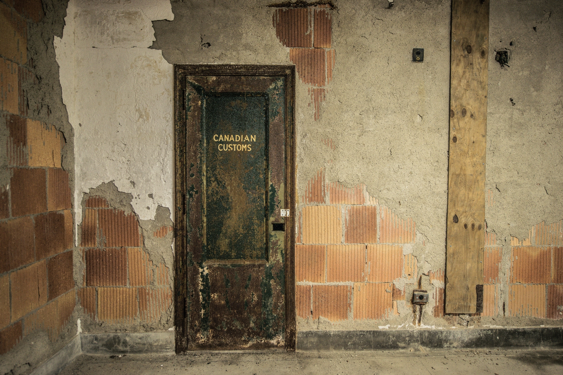Door inside an abandoned building reading "Canadian Customs". The plaster is flaking off the wall, exposing the brickwork underneath.