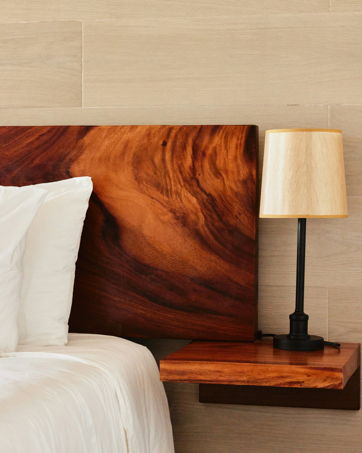 白いシーツの木製ヘッドボード付きベッドの角と、黒とクリーム色のランプのあるサイドテーブル。