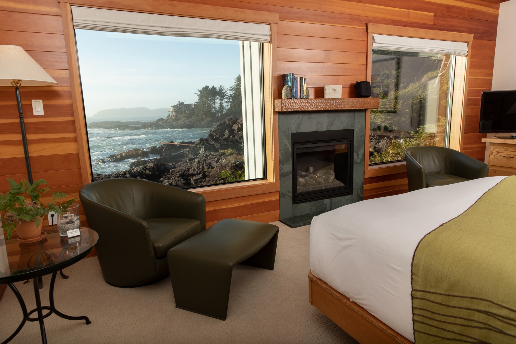 Image of Premier Room in Wickaninnish Inn hotel, a top green hotel in Canada. Hotel bed is made with a green and white blanket on top. Fireplace, table and chair, and view of the Pacific Ccean through the window.