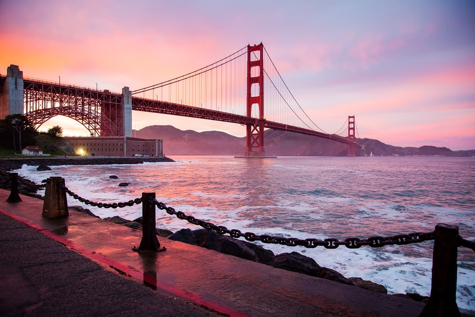 The Golden Gate Bridge spans across the water under a vibrant pink and orange sunset, viewed from a wet shoreline walkway with a black chain railing.