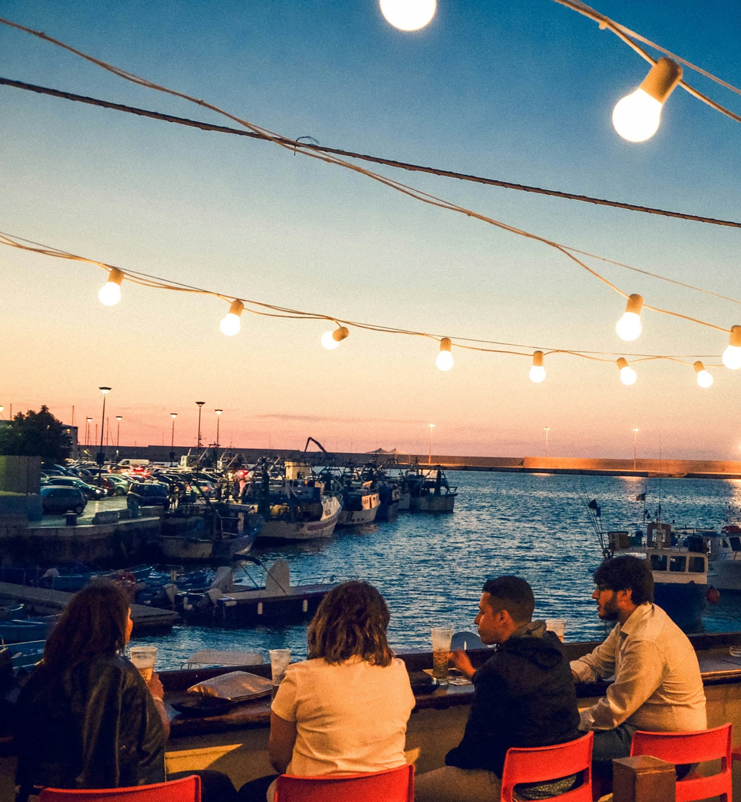 이탈리아 바리에서 어둠이 내리는 항구 카페에 앉아 늘어진 전구 조명 아래 담소를 나누는 사람들