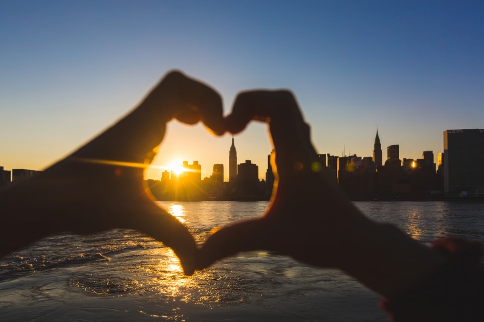 person making a heart shape with their hands with a view of the New York skyline at sunset