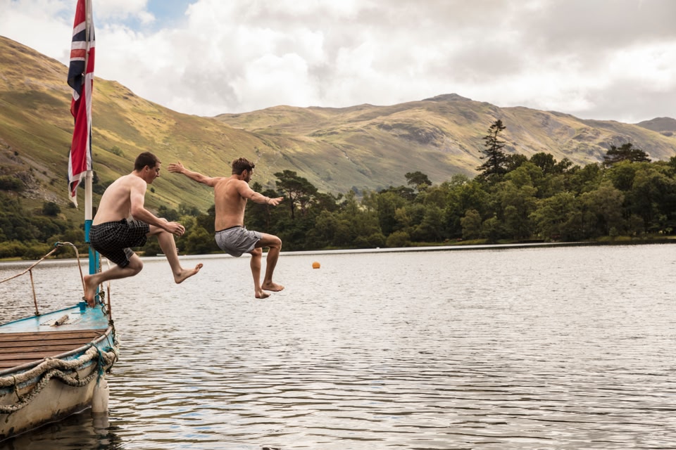 Two men in swimming shorts jumping off a boat into the lake at Ullswater, Lake District, Cumbria.