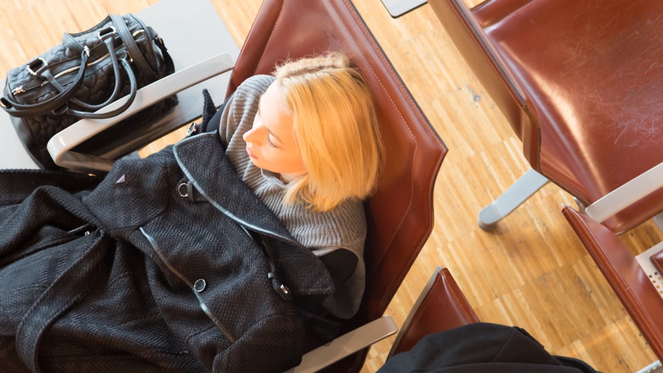 birds eye view of a blond woman sat on a red airport chair with her black coat over her like a blanket 