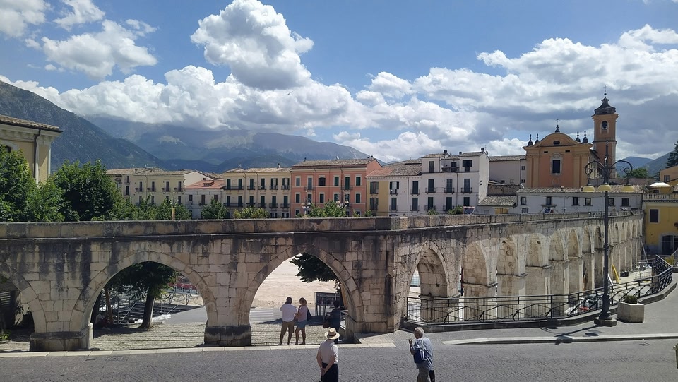 A view of a town dominated by a large, arched stone bridge. The bridge spans a street where people are walking. Behind the bridge, a row of buildings and a church are visible, set against a backdrop of mountains and a cloudy sky.