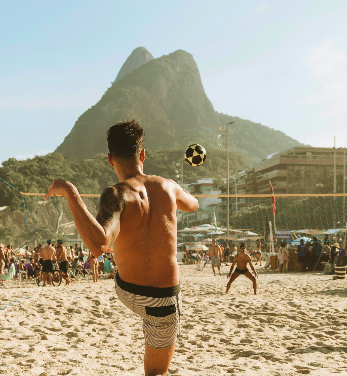 Um homem chuta uma bola de futebol em uma praia movimentada no Rio de Janeiro com o Pão de Açúcar ao longe.