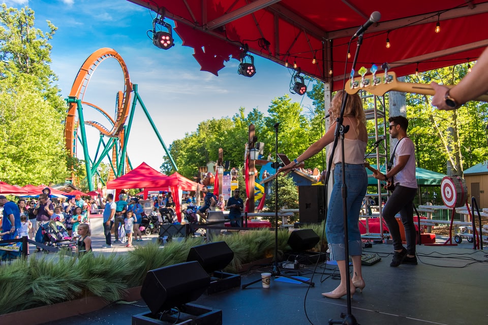 musicians performing on stage with crowds of people and roller coaster in the background