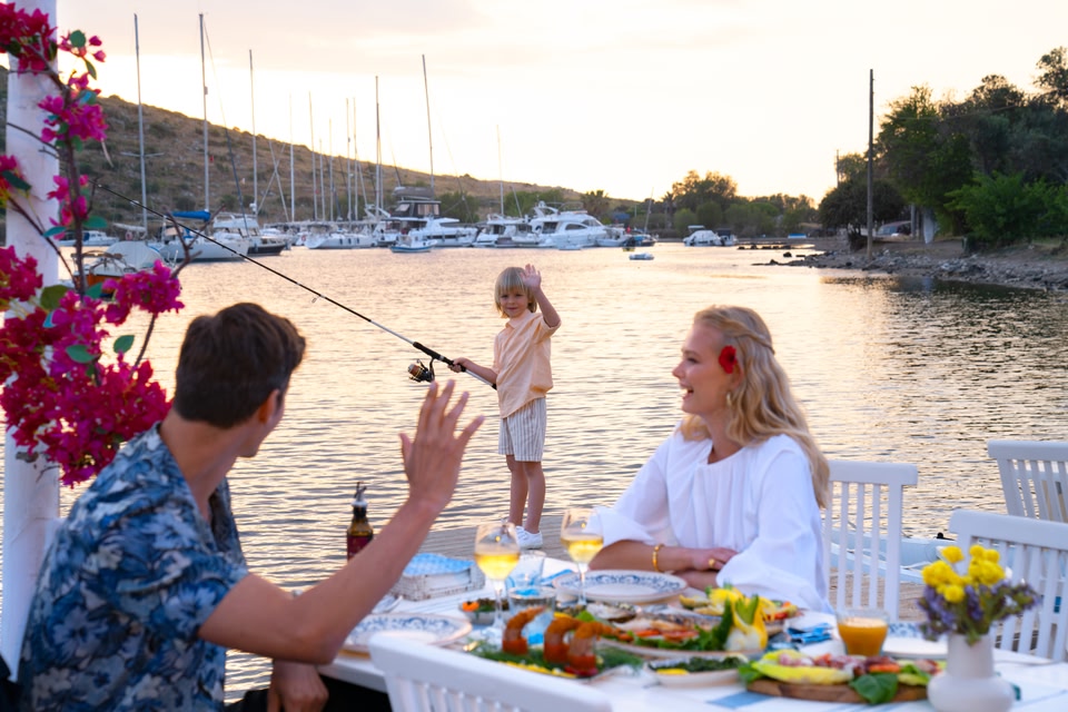 deux personnes partagent un repas devant un port de plaisance avec un enfant pêchant en arrière-plan