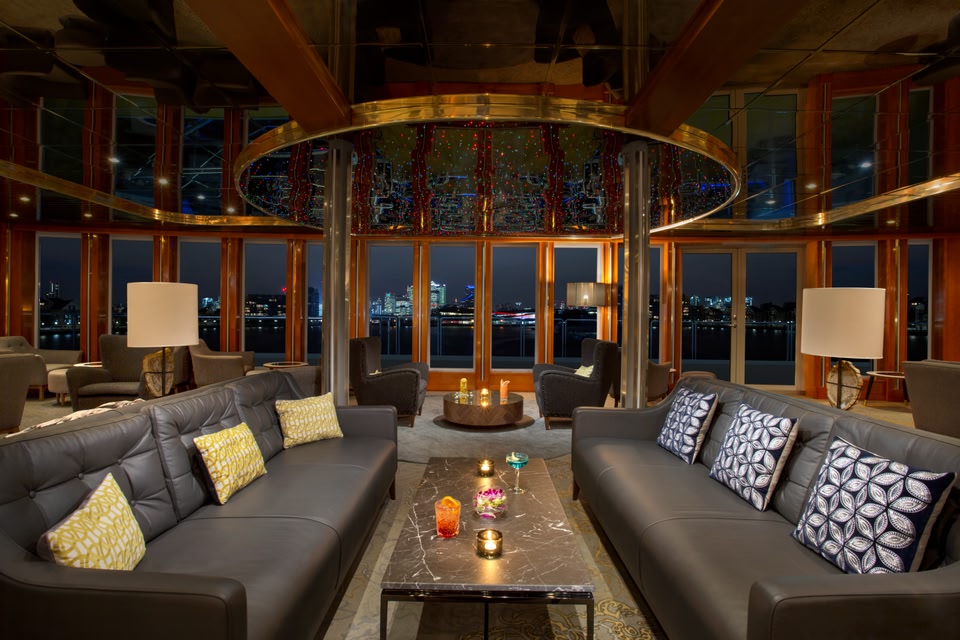 Communal area of Sunborn London Yacht Hotel, with two large leather sofas and a coffee table with cocktails on it. Floor-to-veiling windows show the London skyline at night