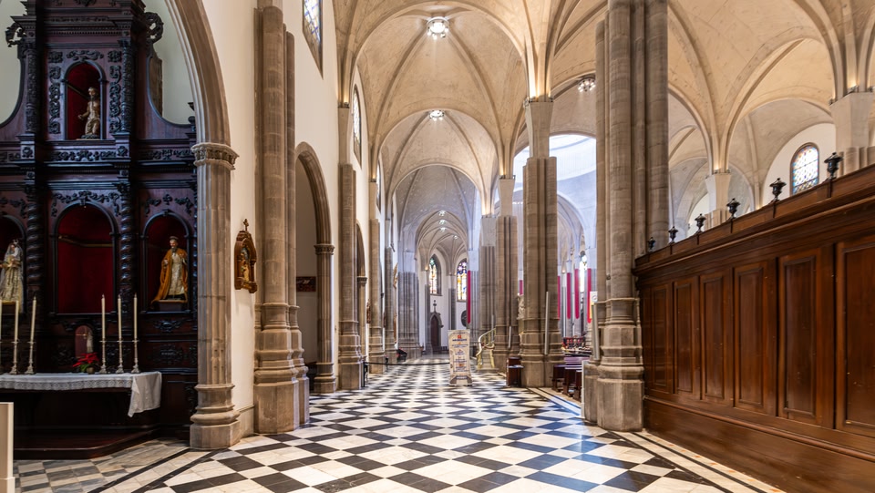 The interior of a grand church with a black and white checkered floor, soaring arches supported by massive columns, and ornate wooden details.